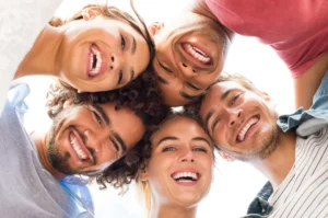 Group of five friends huddled in a circle looking down at the camera with wide, healthy smiles