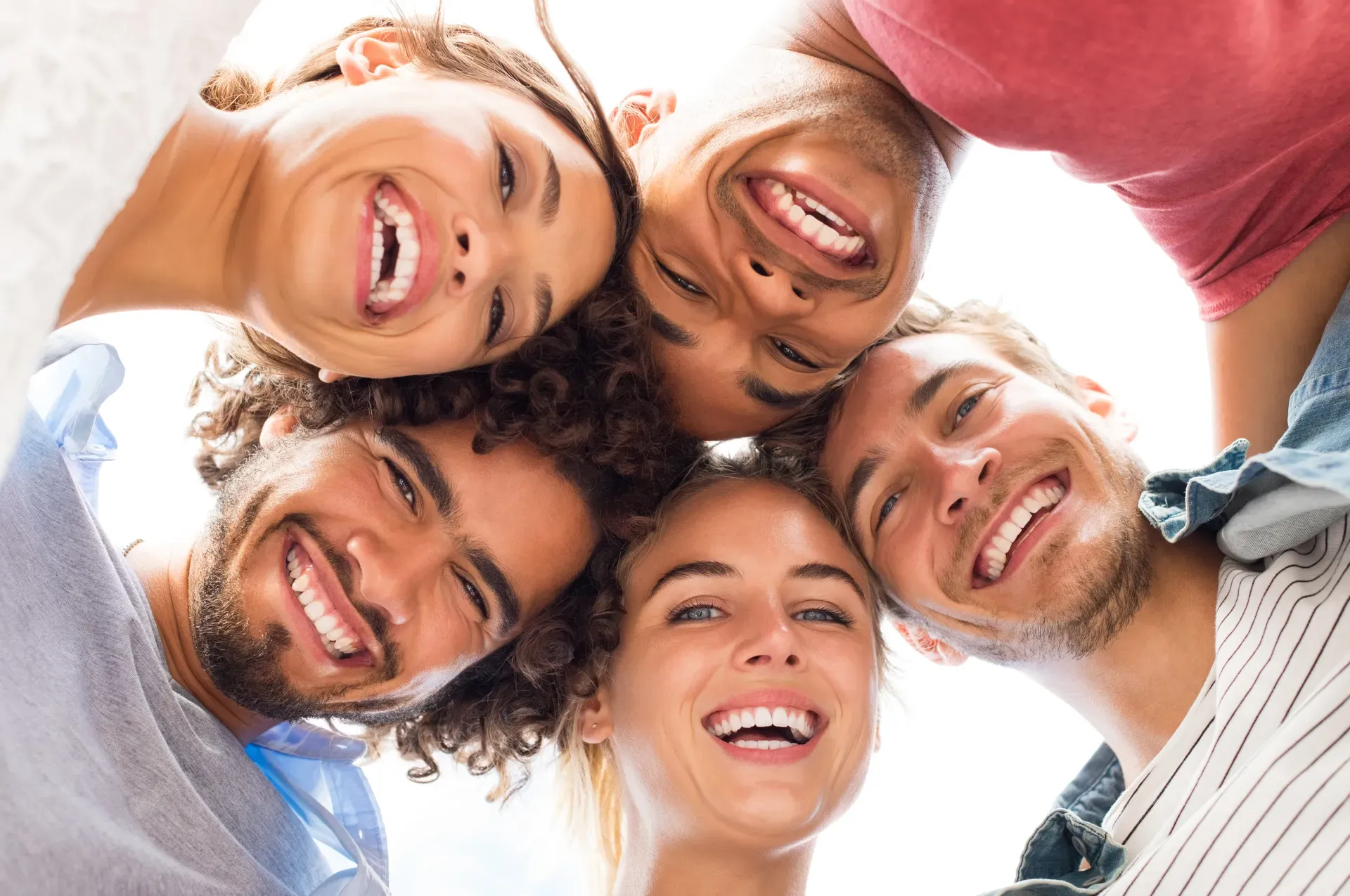 Group of five friends huddled in a circle looking down at the camera with wide, healthy smiles