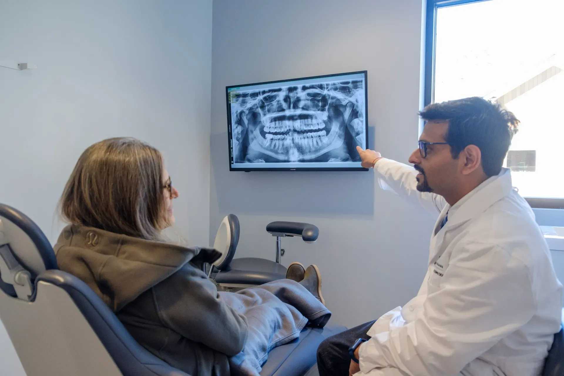 Dentist pointing to a panoramic dental X-ray on a wall-mounted monitor while explaining treatment to a seated female patient in an exam room